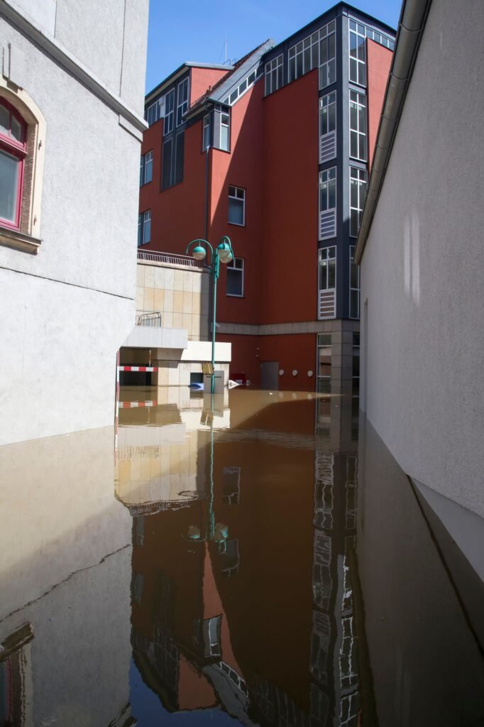 Floodwater covering the ground between city buildings after heavy rain, with a red building reflected in the water